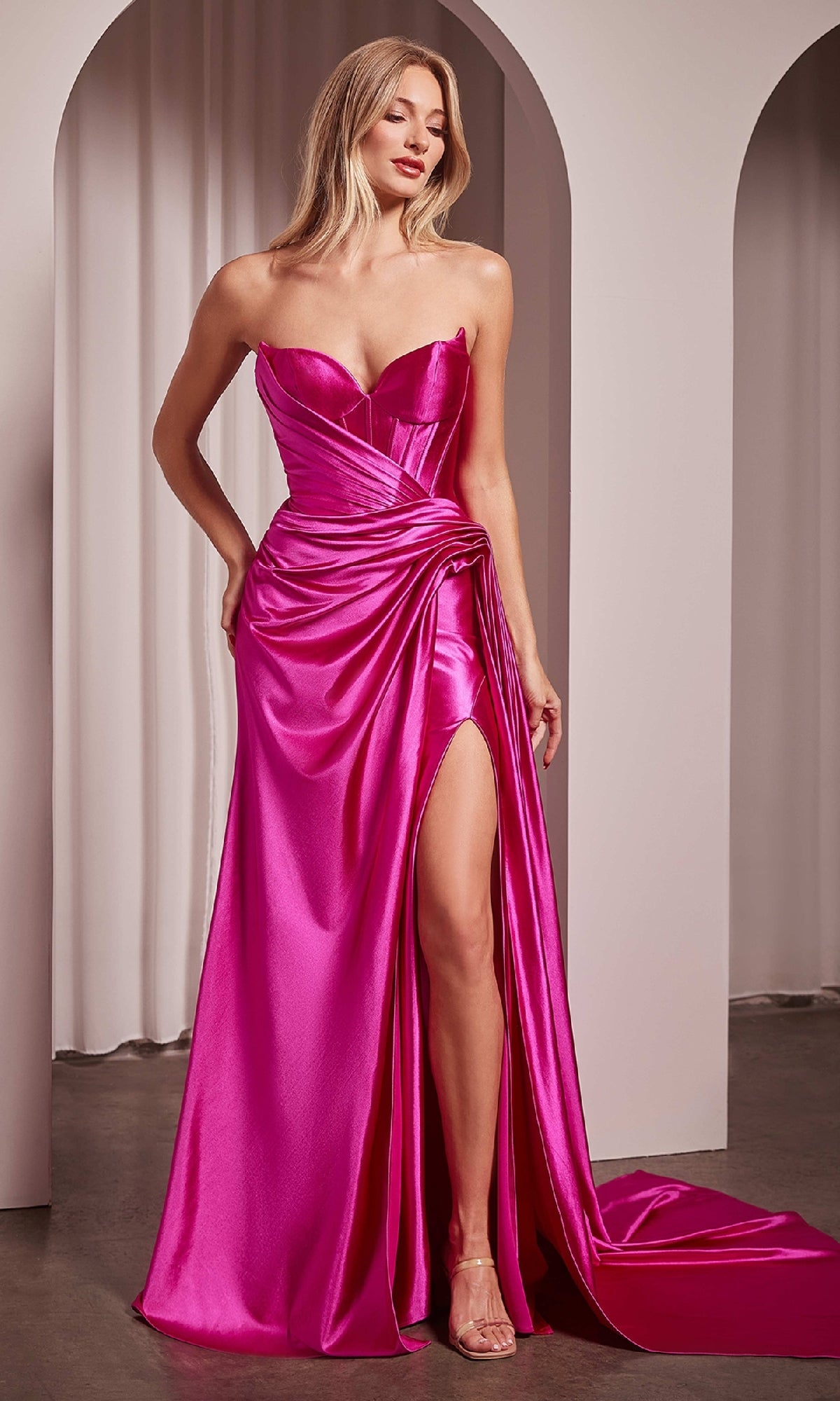 A woman in the Ladivine CD379 Side-Sash Strapless Long Prom Gown, featuring hot pink satin, a sweetheart neckline, draped skirt, and thigh-high slit, stands indoors near white archways.